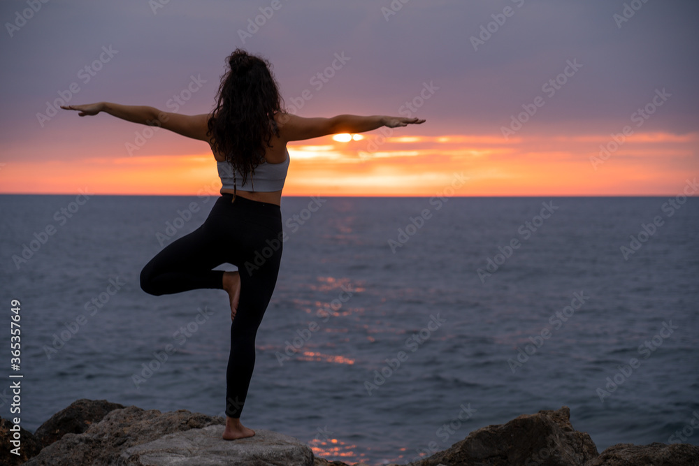 Barefoot young brunette girl practicing yoga in front of the mediterranean sea while the sun is almost on the horizon and shining with an orange light while standing on a rock