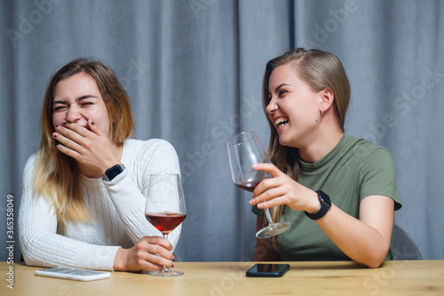 two girls of European appearance with blond hair are sitting at the table  drinking wine and laughing  relaxing at home  alcohol