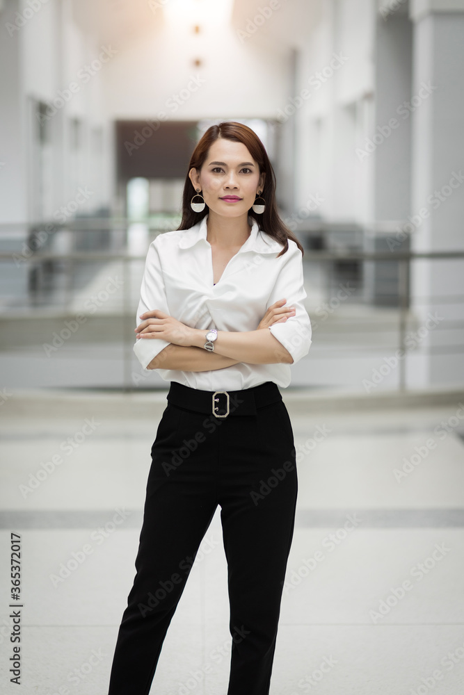 Portrait of a cheerful middle-age businesswoman in business suit stands in the company building with confidence arms crossed. Modern business woman in the office with copy space. Business stock photo. Portrait of a cheerful middle-age businesswoman in business suit stands in the company building with confidence arms crossed. Modern business woman in the office with copy space. Business stock photo.