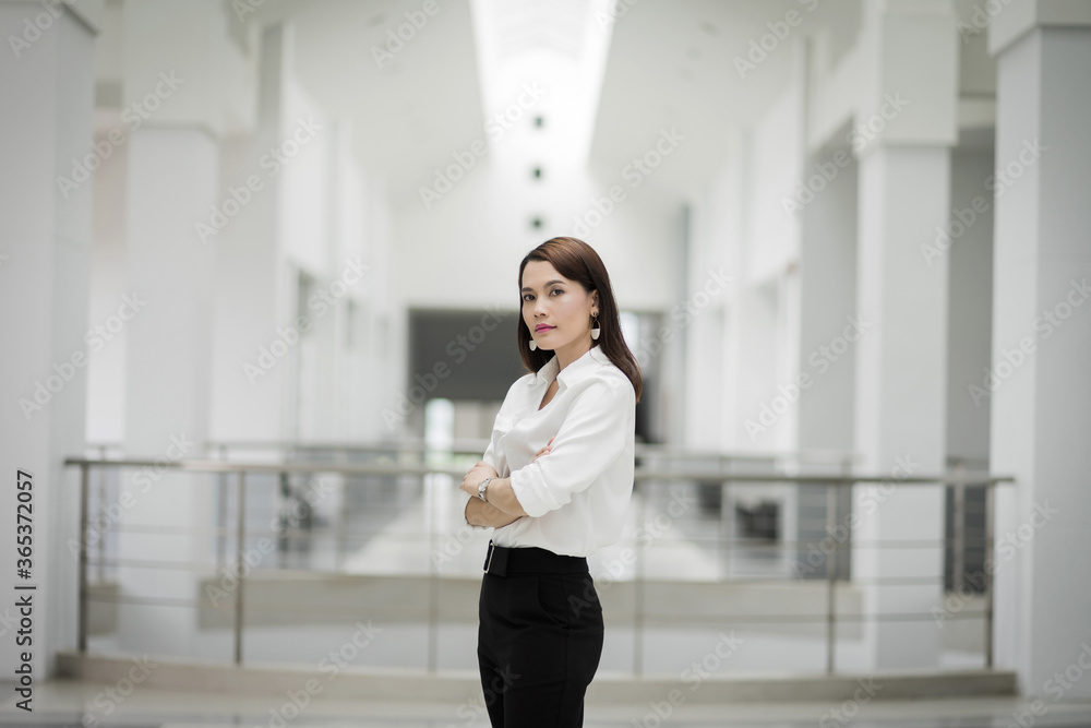 Portrait of a cheerful middle-age businesswoman in business suit stands in the company building with confidence arms crossed. Modern business woman in the office with copy space. Business stock photo. Portrait of a cheerful middle-age businesswoman in business suit stands in the company building with confidence arms crossed. Modern business woman in the office with copy space. Business stock photo.