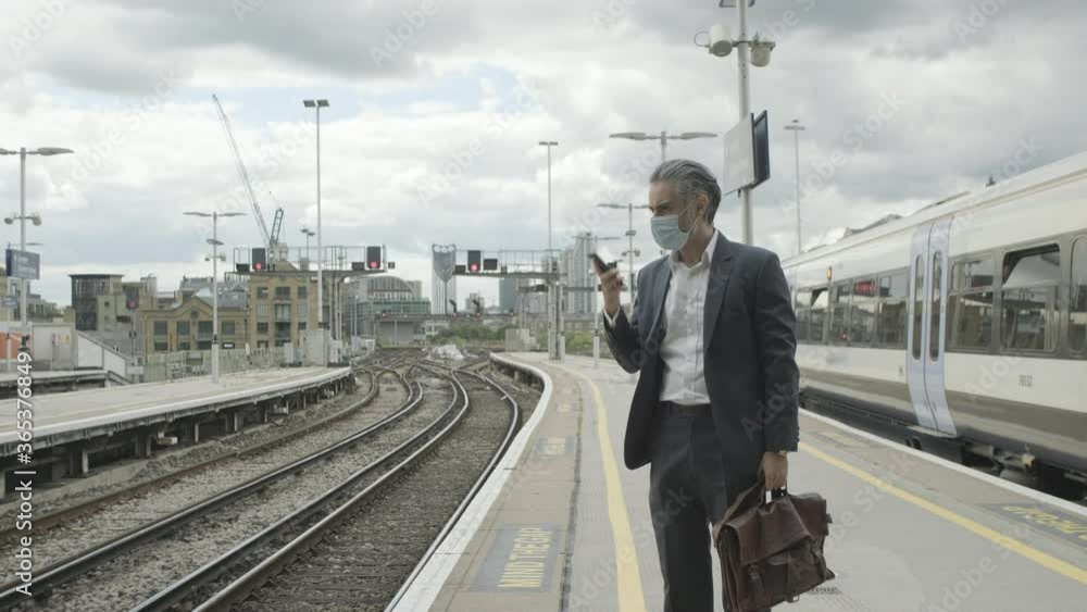 Mature business man wearing face mask standing on train station platform talking on smart phone Mature business man wearing face mask standing on train station platform talking on smart phone