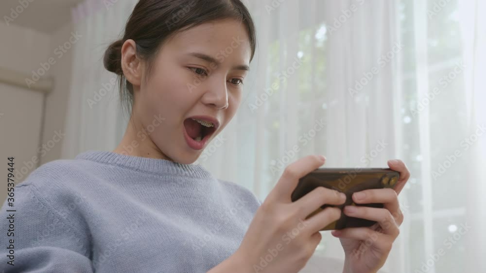 excited young Asian woman playing games on smartphone in the living room at home. excited young Asian woman playing games on smartphone in the living room at home.
