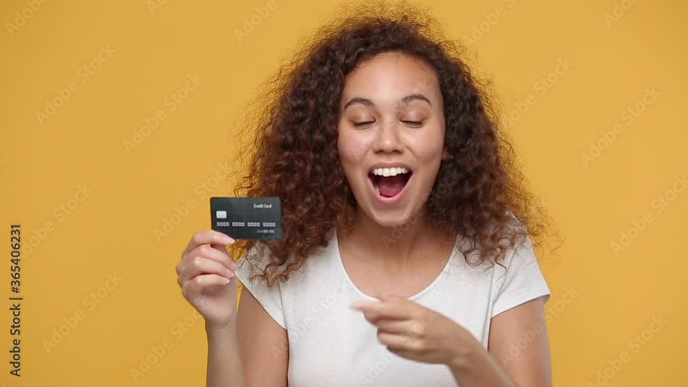 Young happy african american girl 20s years old in white t-shirt isolated on yellow background studio. People lifestyle concept. Looking camera hold in hand bank credit card showing thumbs up gesture