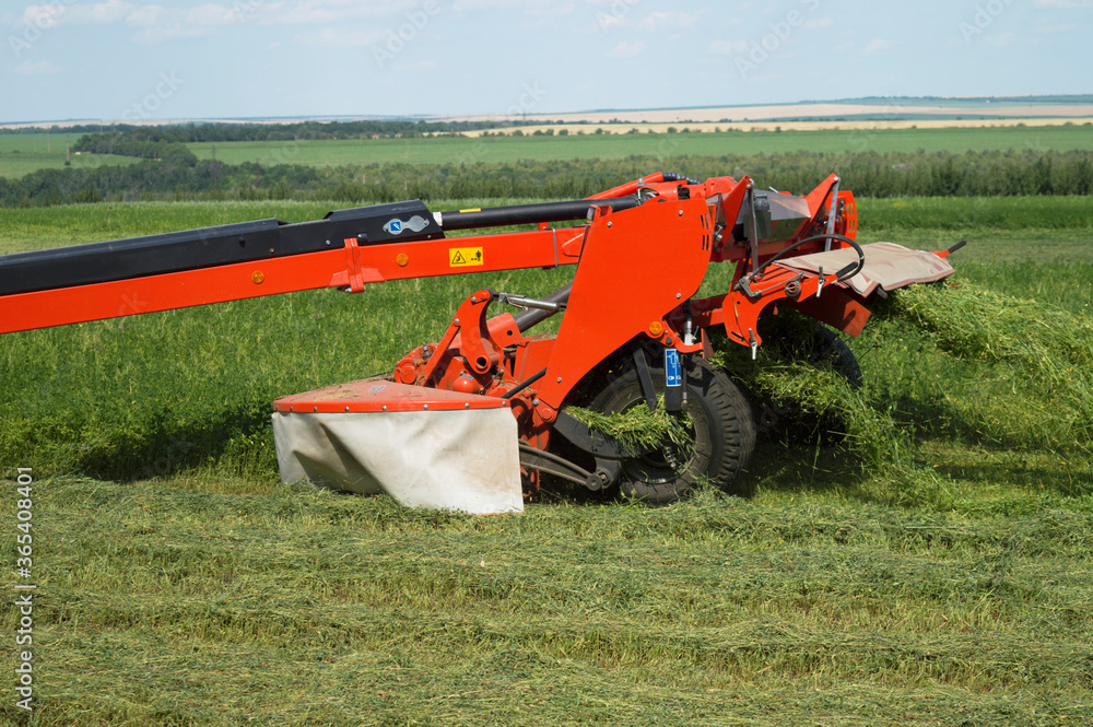 Red tractor trailer cutting grass on the field. The process of mowing clover. Summer work on an agricultural farm. Harvesting equipment. Red tractor trailer cutting grass on the field. The process of mowing clover. Summer work on an agricultural farm. Harvesting equipment.