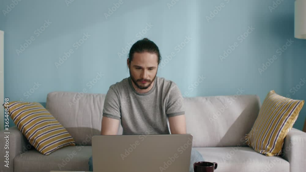 Young man, freelancer, works at the computer, sitting on sofa at home, enjoying his working day and home workplace, slow motion. Young man, freelancer, works at the computer, sitting on sofa at home, enjoying his working day and home workplace, slow motion.