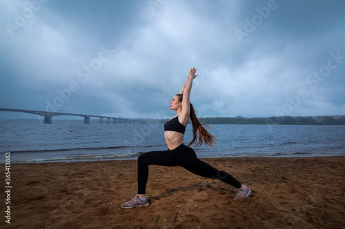 Young woman doing yoga outside. bad weather early morning. Overcome self motivation. A strong girl stands in the pose of a warrior asana doing exercises. workout harmony zen balance. Cloudy sky beach