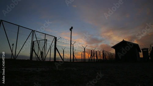 Time-lapse of a sunset  and clouds moving with prison in background silhouette with barbed wire and fence. 