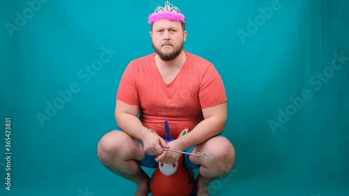 handsome bearded freaky man in a pink T-shirt with a deadema on his head is saddened riding a unicorn with a magic wand in his hand. A funny wizard joke to make and fulfill a wish. photo