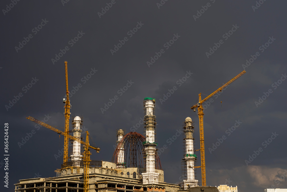 Fototapeta premium Construction of a mosque. Thunderclouds. The unfinished white mosque shines amid storm clouds. Clouds. Rainbow and Mosque. Crane. Concrete blocks and metal frame. Major construction in the city