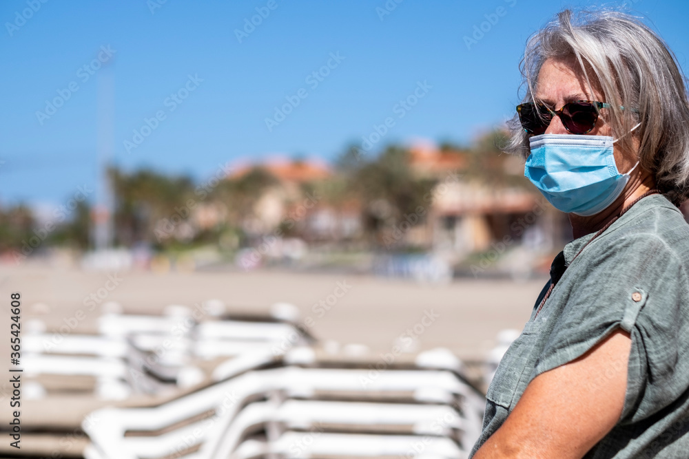 Senior adult woman wearing a protective mask looks at deserted beach with closed umbrellas and  beach beds -  lockdown due to the Covid-19 coronavirus.