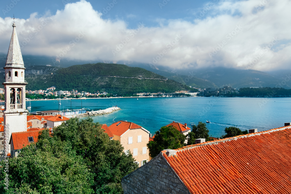 Church tower and red roofs of ancient town Budva and Adriatic sea coast. Medieval architecture.White clouds blue sky and aqua green sea water background