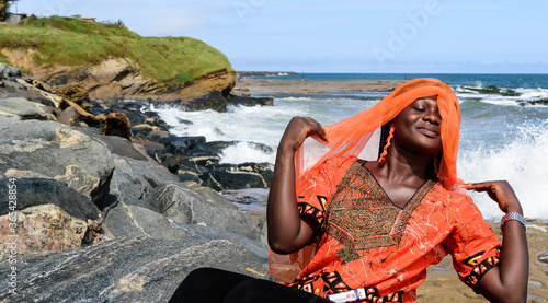 African woman from Ghana with orange dress on the rocks by the sea in Takoradi Ghana West Africa photo