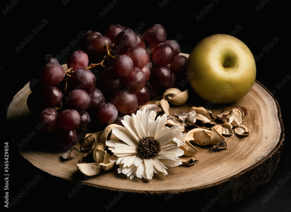 Still life of grapes and apple with a flower in the middle and dried leaves on top of a piece of wood and black background