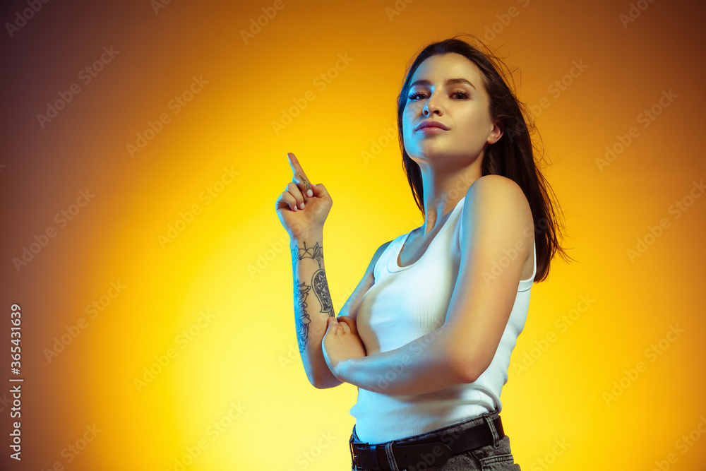 Pointing at side. Portrait of young caucasian woman isolated on gradient yellow studio background. Stylish, trendy look. Beautiful model in casual. Human emotions, facial expression, sales, ad concept Pointing at side. Portrait of young caucasian woman isolated on gradient yellow studio background. Stylish, trendy look. Beautiful model in casual. Human emotions, facial expression, sales, ad concept