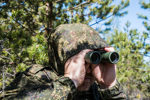 military scout or spy watches through binoculars from the depths of the forest