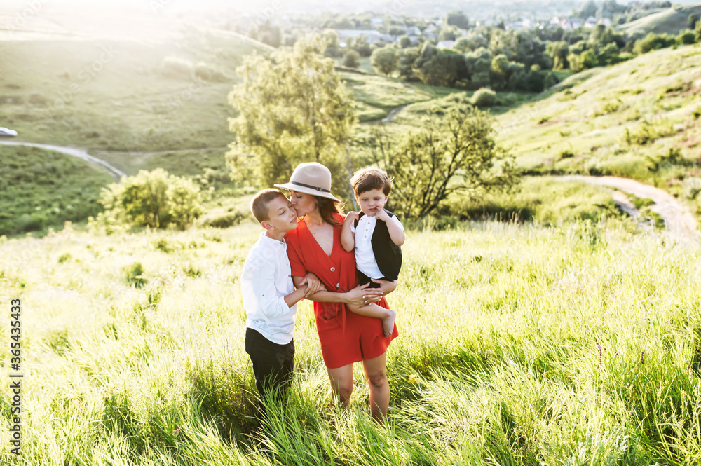 Young woman with two little male kids stands in the middle of a green field. She kisses the older one while holding the younger on her hands. Gentle happy family outside Young woman with two little male kids stands in the middle of a green field. She kisses the older one while holding the younger on her hands. Gentle happy family outside