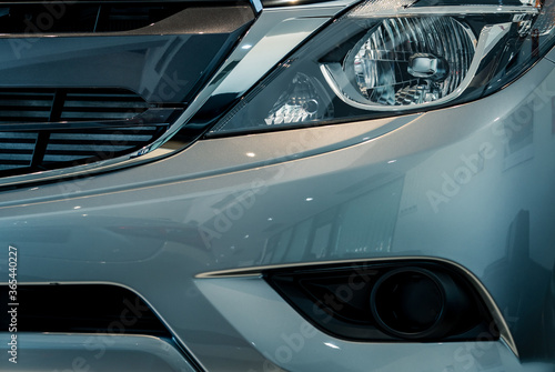 Closeup headlamp of gray SUV car parked in modern showroom. Shiny new car with reflection of modern showroom. Automotive industry. Electric car technology. Automobile headlight. Car dealership concept © Artinun