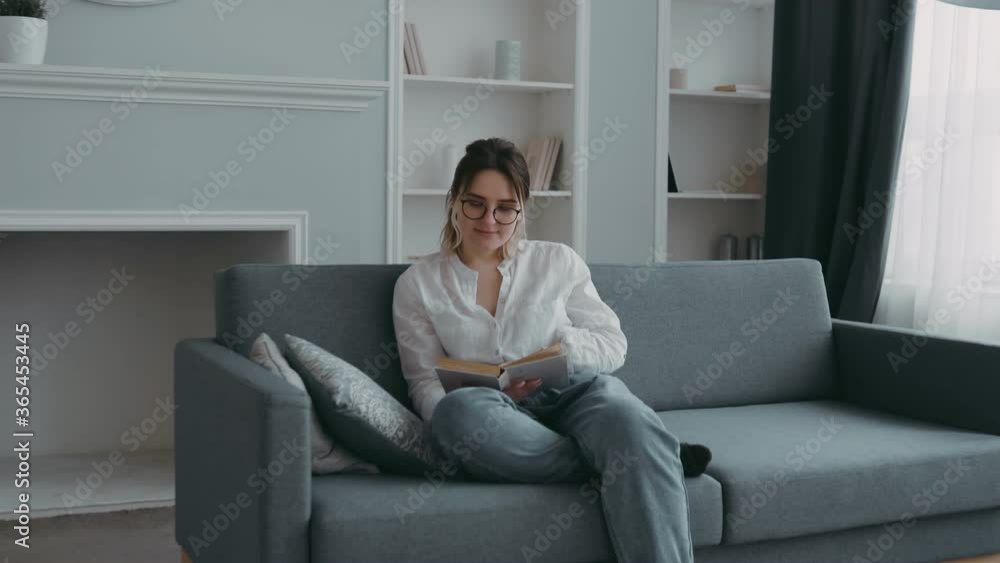 Young beautiful smart girl with glasses sitting on sofa in front of window in modern living room reading book. Close-up portrait of happy, smiling woman student. Concept of home rest and education. Young beautiful smart girl with glasses sitting on sofa in front of window in modern living room reading book. Close-up portrait of happy, smiling woman student. Concept of home rest and education.