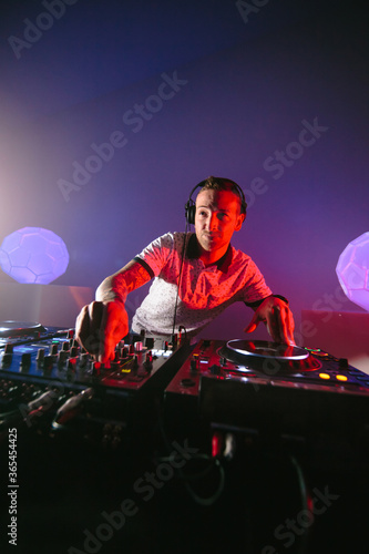 Fotografie Vertical shot of a young Caucasian Techno male DJ with headphones with his mixer board playing music in a purple background