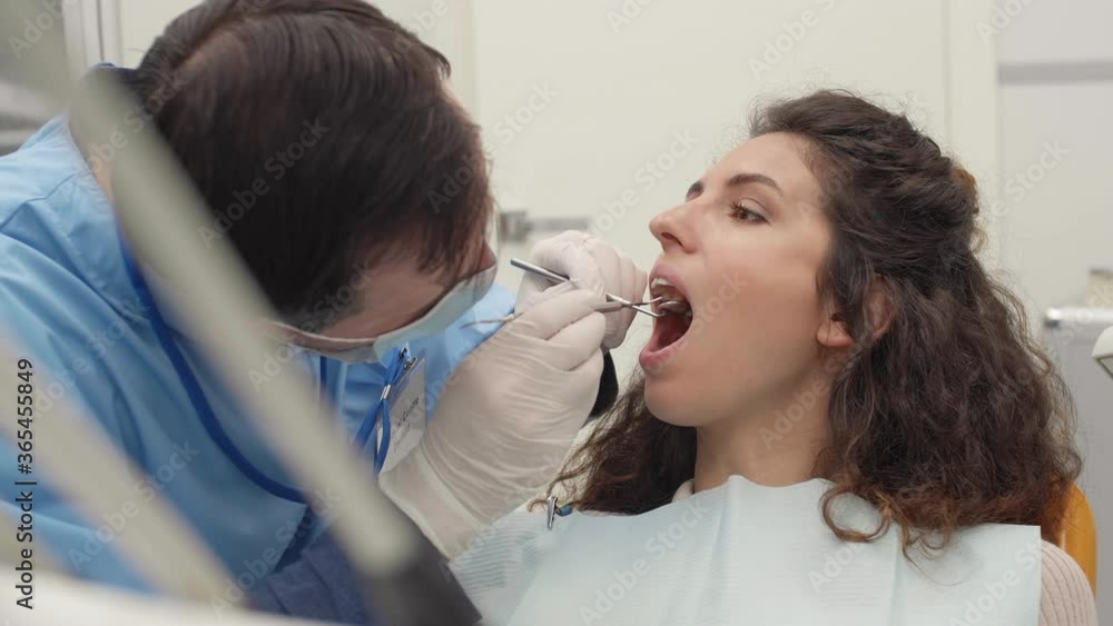 Close up of male dentist wearing medical overalls, mask and gloves holding special tools and checking teeth health of woman lying on dental chair Close up of male dentist wearing medical overalls, mask and gloves holding special tools and checking teeth health of woman lying on dental chair