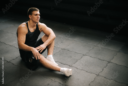 Strong muscular man with perfect beautiful body wearing sportswear sitting on the gym floor with weight from the barbell after heavy workout training in dark modern gym.