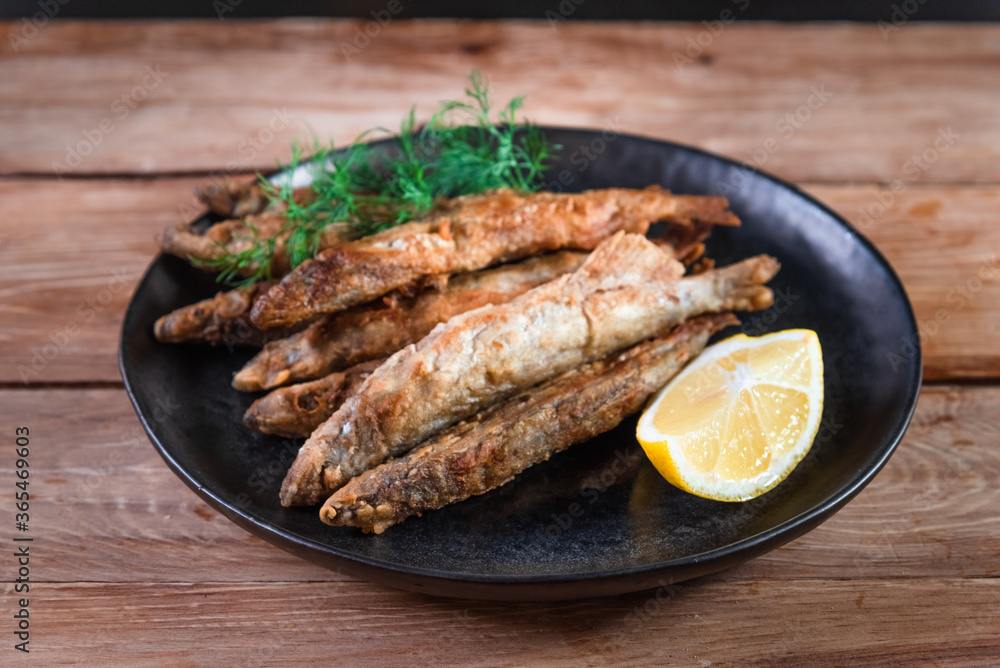 Appetizing fried capelin on a black plate stands on a wooden surface. Simple rustic food concept Appetizing fried capelin on a black plate stands on a wooden surface. Simple rustic food concept