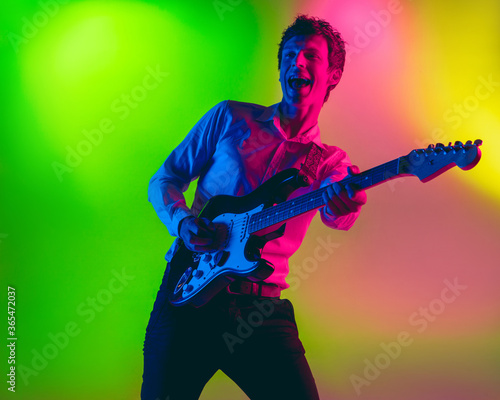 Talented. Young caucasian inspired and expressive musician, guitarist performing on multicolored background in neon. Concept of music, hobby, festival, art. Joyful artist, colorful, bright portrait.