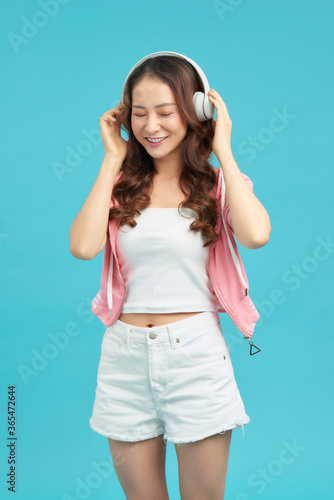 Cheerful stylish young woman standing isolated over blue background, listening to music with headphones, dancing