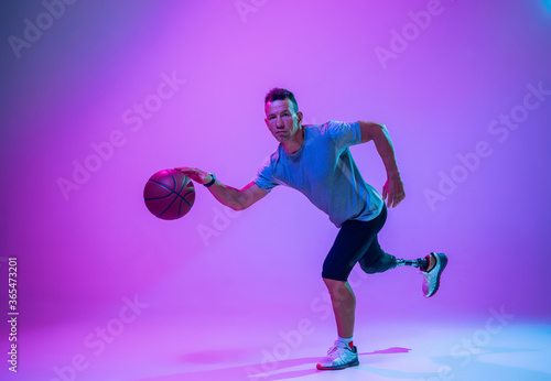 Athlete with disabilities or amputee on gradient studio background in neon. Professional male basketball player with leg prosthesis training in studio. Disabled sport and healthy lifestyle concept.