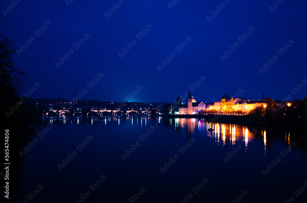 An early evening in Prague during autumn with incandescent lights illuminating the city and reflections over the Vltava river.