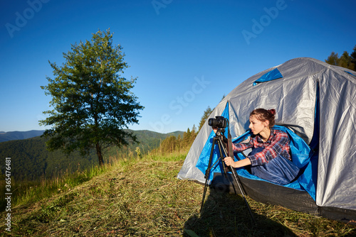 Summer morning camping in the mountains. Young female photographer sitting in tourist tent, taking photo, using photo camera and tripod. On background blue sky and big tree.