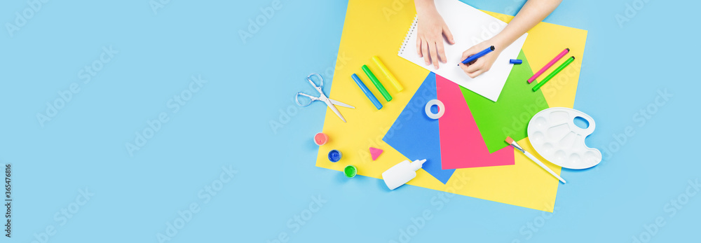 Obraz premium Hands drawing over color paper, scissors, pencils paint and various school stationery on blue yellow background. Flat lay with copy space for back to school or education and craft concept banner