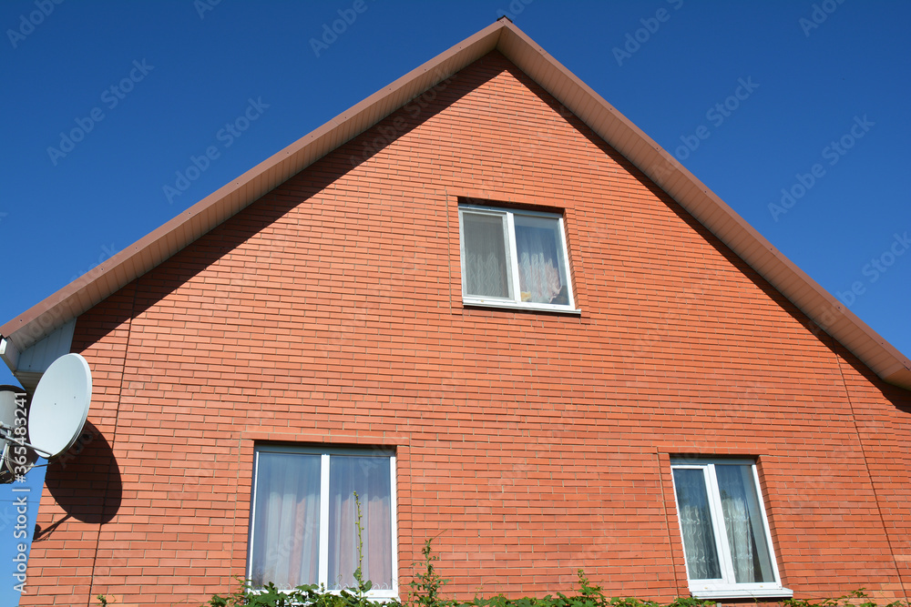 The facade of a brick house with a mansard window, satellite antenna, metal roof, plastic fascia and soffit against blue sky.