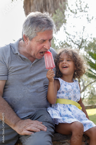 Bild auf Leinwand Nonno bianco mangia un ghiacciolo  con la nipotina Brasiliana seduti in un parco