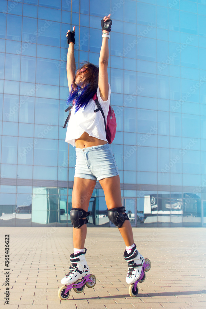 Young beautiful fit girl with blue hair, roller skates, with backpack and in protection jumping to the sky against the background of blue glass building. Summer sports lifestyle. Informal Roller Girl