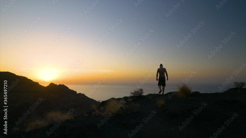 Silhouette of Man standing in Cliff next to ocean with Beautiful Sunset in Horizon then sits down to look at Sunset Silhouette of Man standing in Cliff next to ocean with Beautiful Sunset in Horizon then sits down to look at Sunset