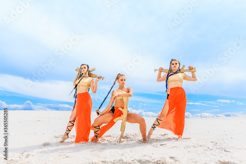 Girls dancers in red skirts and with golden machines in their hands on the beach. White sand and blue sky.