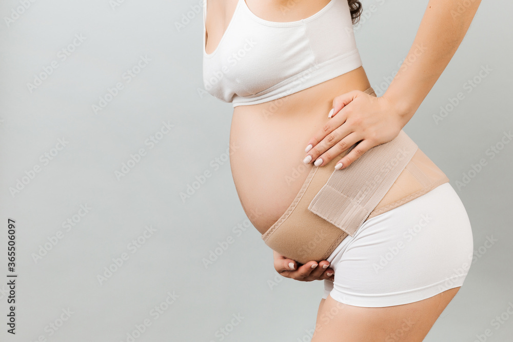 Close up of supporting bandage against backache on pregnant woman in  underwear at gray background with copy space. Mother is suffering from pain  in the back. Orthopedic abdominal support belt concept Photos
