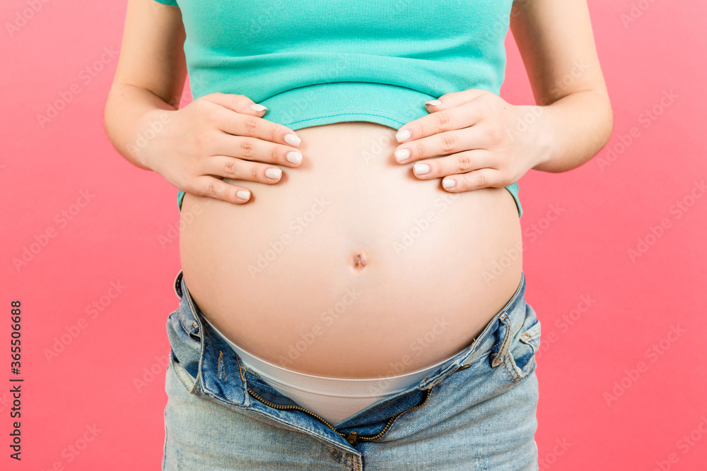 Close up of expecting mother in unzipped jeans showing her naked pregnant belly at colorful background with copy space. Pregnancy concept Close up of expecting mother in unzipped jeans showing her naked pregnant belly at colorful background with copy space. Pregnancy concept