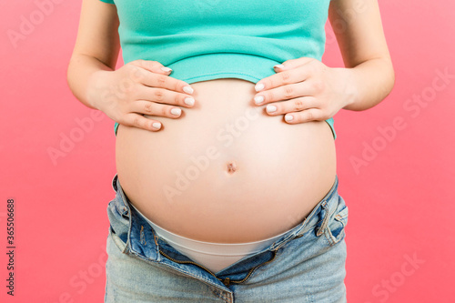 Close up of expecting mother in unzipped jeans showing her naked pregnant belly at colorful background with copy space. Pregnancy concept © sosiukin