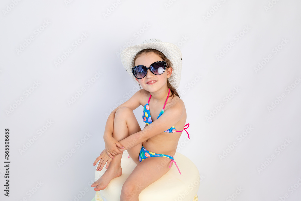 Cute little girl in black sunglasses and a blue bikini is sitting on the floor , turning sideways to the camera-Isolated on white background