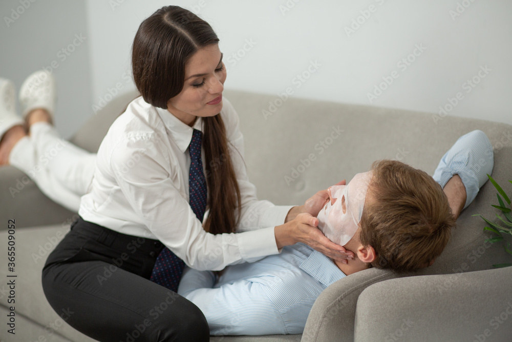 Obraz premium Woman putting face mask on her husband. Caring young lady wearing formal clothing putting sheet mask on her boyfriends face while he lays on couch.