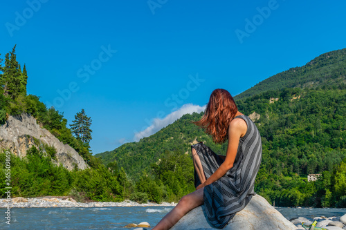 An unrecognizable relaxed young Caucasian redhead woman sitting on a boulder with a beer bottle next to the Var river in the French Alps enjoying summer sunshine