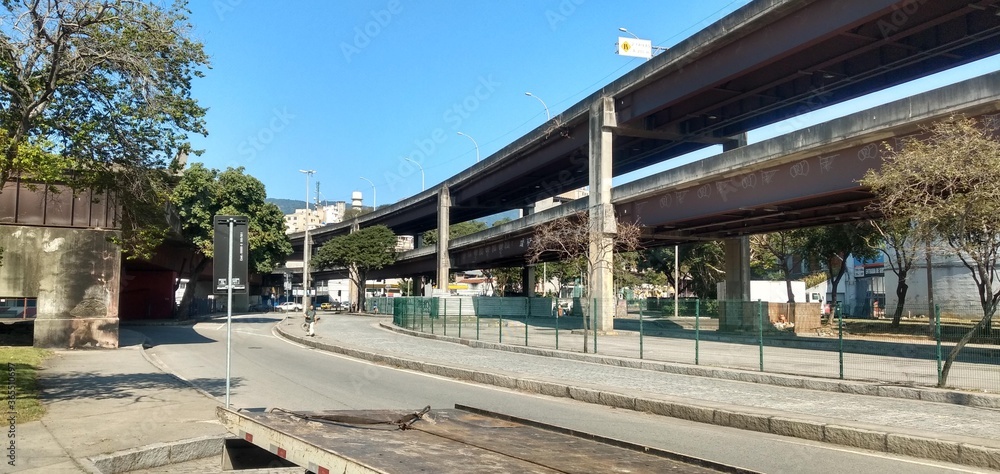 Street in a neighborhood in the north of Rio de Janeiro, shows the street with its trees, right next to it a viaduct with graffiti.