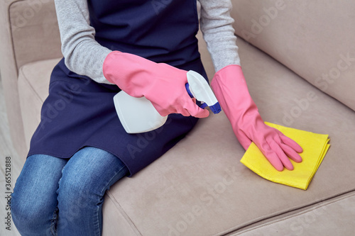 A girl in gloves and an apron cleans upholstered furniture in the living room.