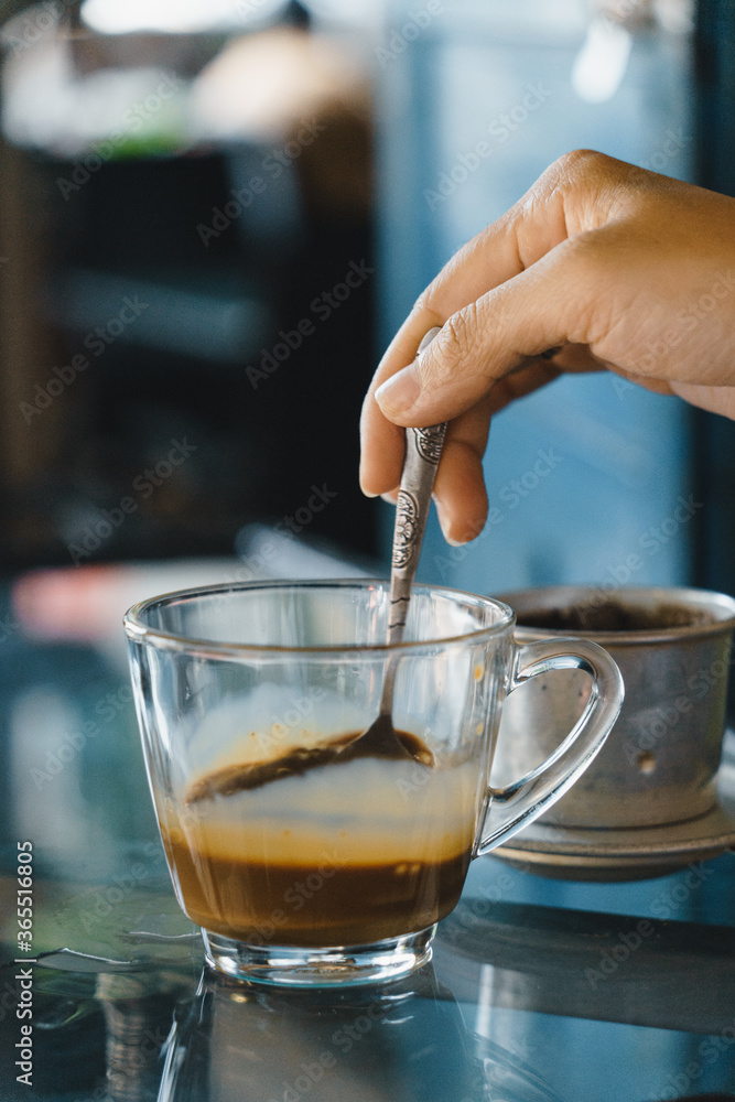 close-up hands of a woman stirring coffee, Hot milk coffee dripping in Vietnam style, pouring water over ground coffee contained in Vietnamese Phin Filter on table. Selective Focus. close-up hands of a woman stirring coffee, Hot milk coffee dripping in Vietnam style, pouring water over ground coffee contained in Vietnamese Phin Filter on table. Selective Focus.
