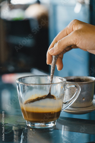 close-up hands of a woman stirring coffee, Hot milk coffee dripping in Vietnam style, pouring water over ground coffee contained in Vietnamese Phin Filter on table. Selective Focus.