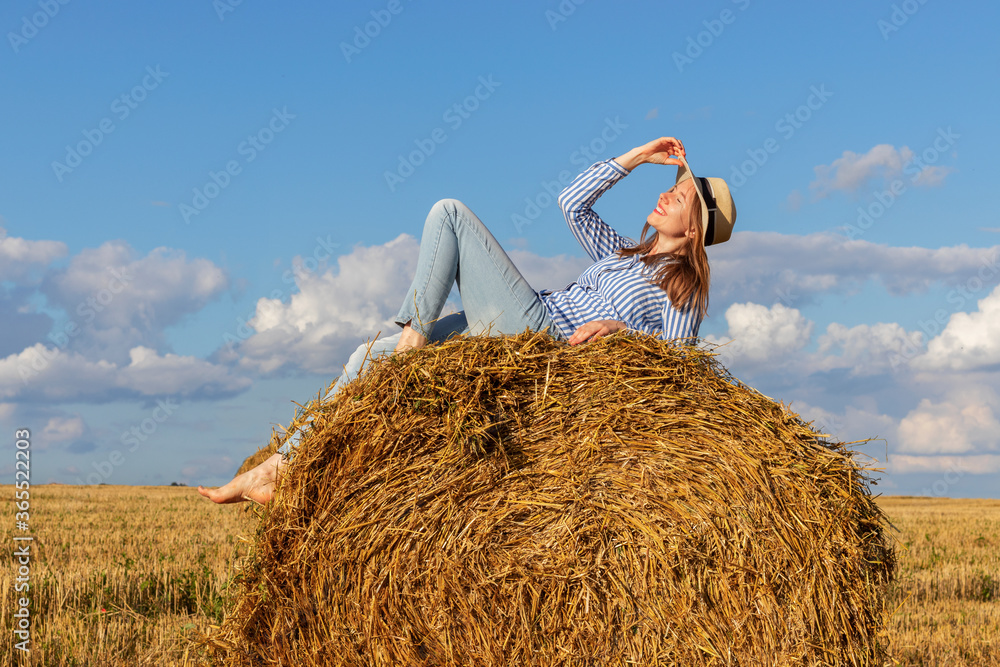 A young woman in blue jeans, shirt and a straw hat lies on a haystack. Female portrait in field in countryside. A young woman in blue jeans, shirt and a straw hat lies on a haystack. Female portrait in field in countryside.