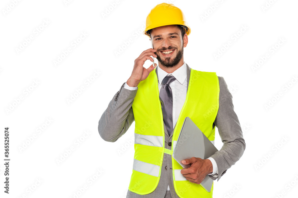 Handsome engineer smiling at camera while talking on smartphone and holding laptop isolated on white