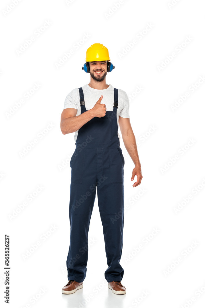 Handsome builder in workwear and hardhat smiling and showing thumb up gesture on white background